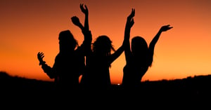 Three women at sunset with hands raised.