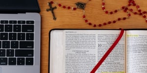 An open bible, rosary, and laptop on a table.