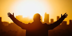 Man with arms open wide in front of a city setting sun. 