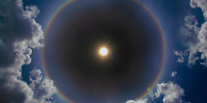 sun in the sky surrounded by clouds and a ring of rainbow light